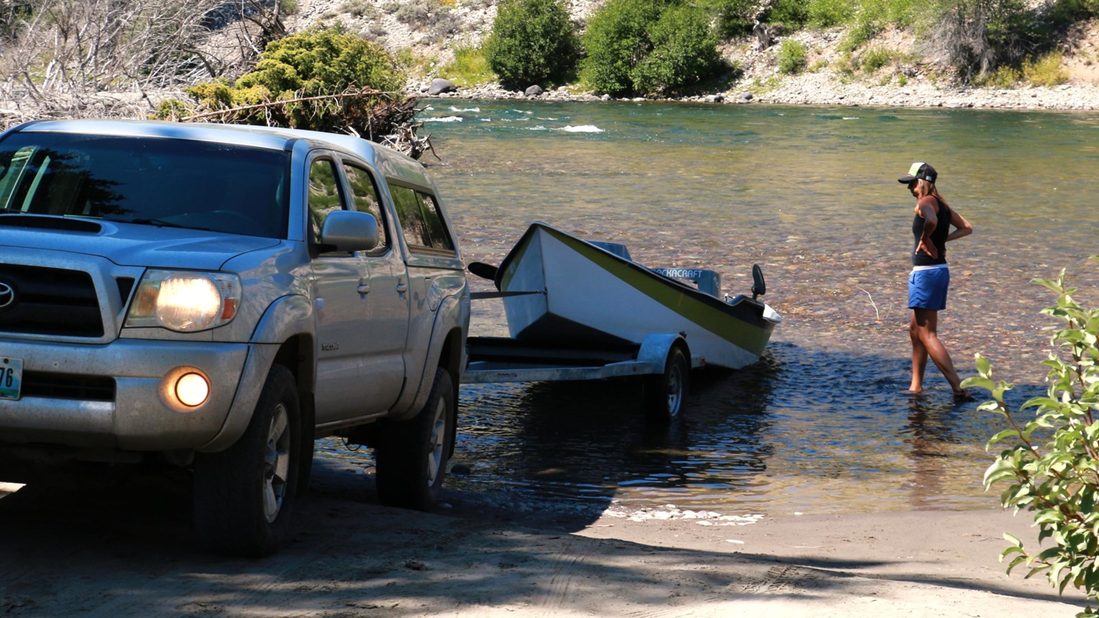 A truck unloads a boat into the river while a person watches.