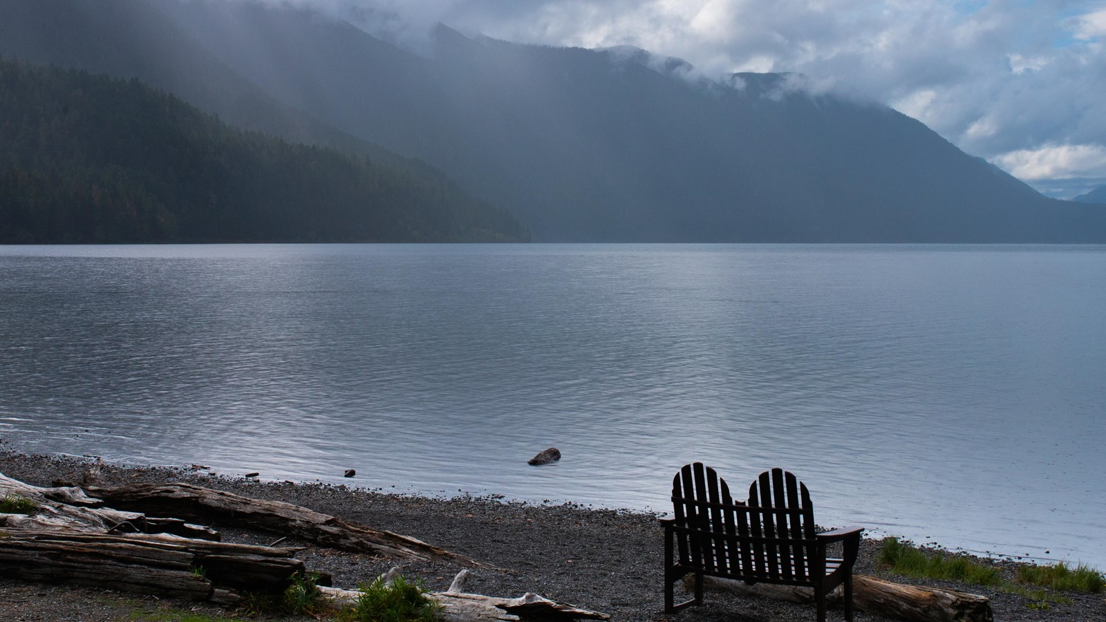 A wooden love seat next to a peaceful lake.