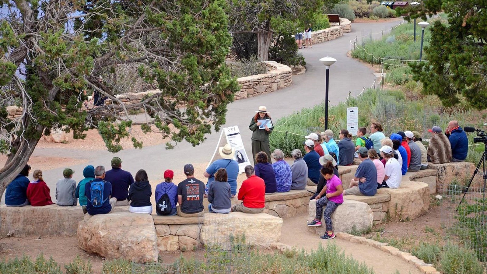 Alongside a paved footpath, 35 people are sitting on stone benches listening to a park ranger talk.