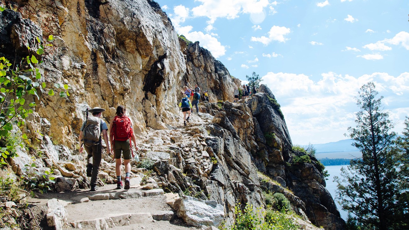 Hikers walk up a rocky trail with a cliff on the right.