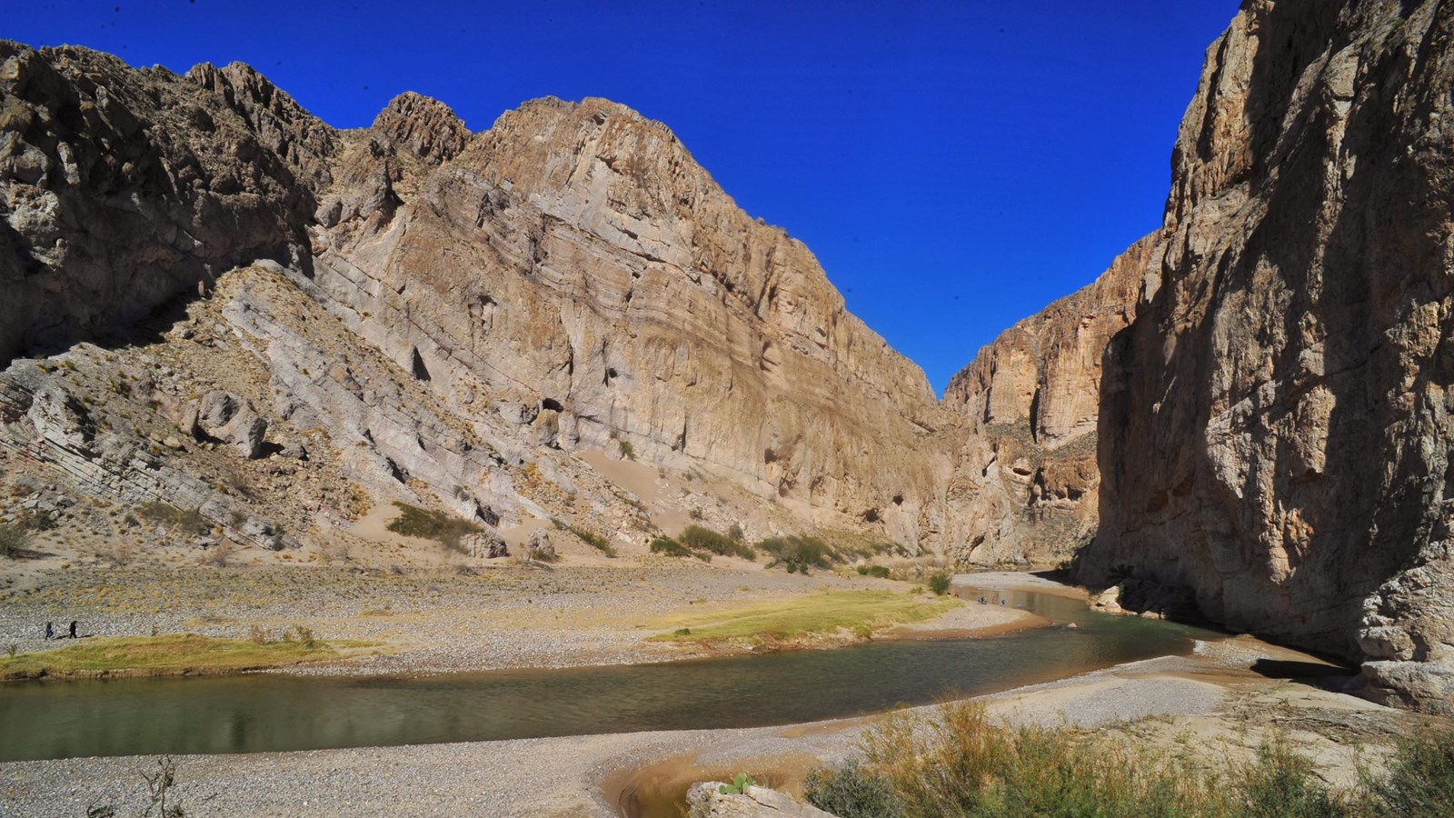 A river flows through high walls of limestone, while hikers walk along the sandy banks.
