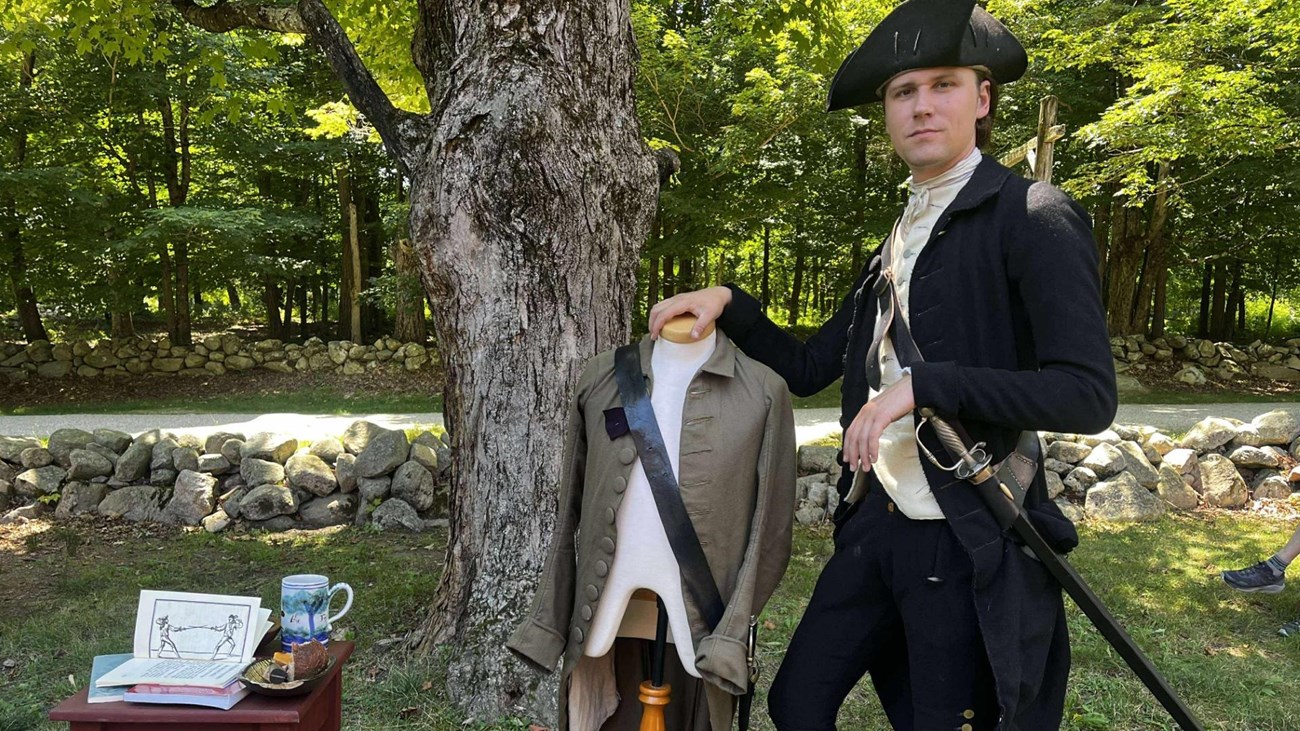 A colonial man in black clothing holds a sword and stands in front of a display about fencing