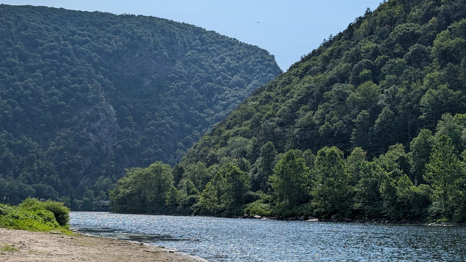 Two mountains slope down to meet the Delaware River. Green foliage covers the mountains.