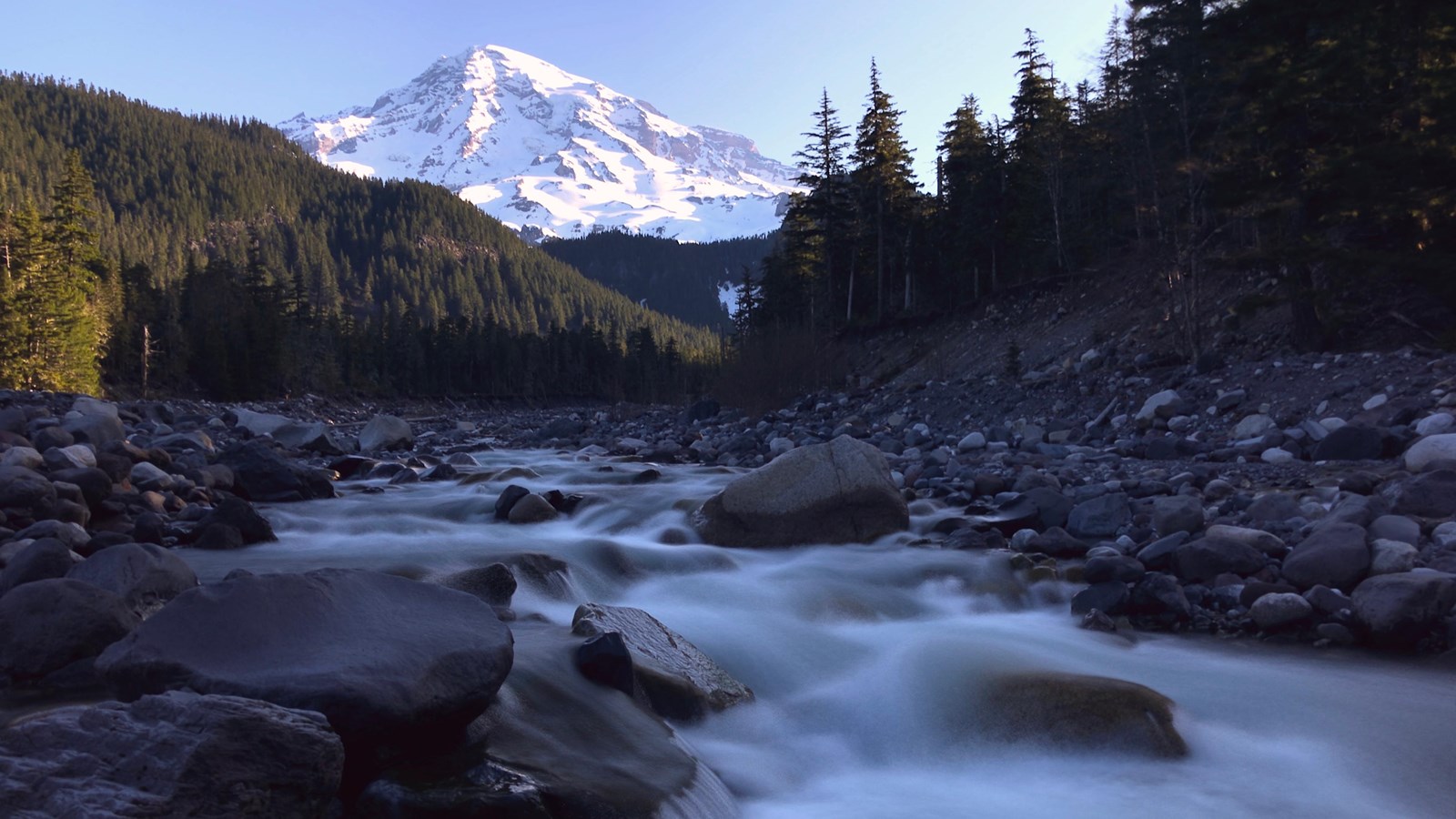 A river flows through a rocky riverbed in a valley on the slopes of a glaciated mountain.