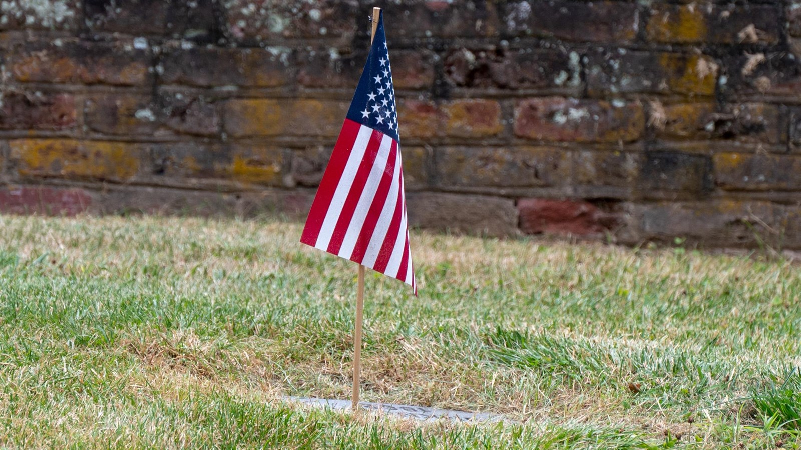 A small United States flag marking a flat stone grave on a lawn in front of a brick wall.