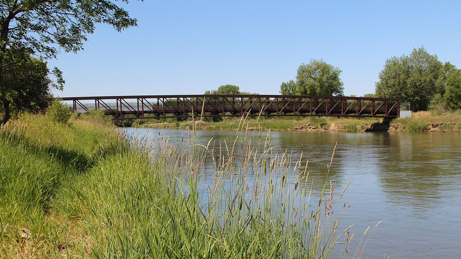 Tall grasses line the riverbank beside a slender brown bridge over the reflective North Platte.