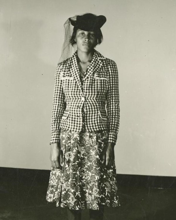 Black & white photo of a Black woman wearing a floral dress, jacket and hat.