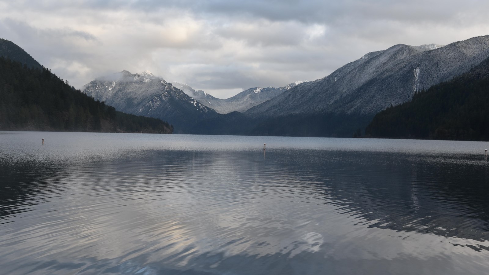 A serene lake under cloudy skies, with hills and snowy mountains in the distance.