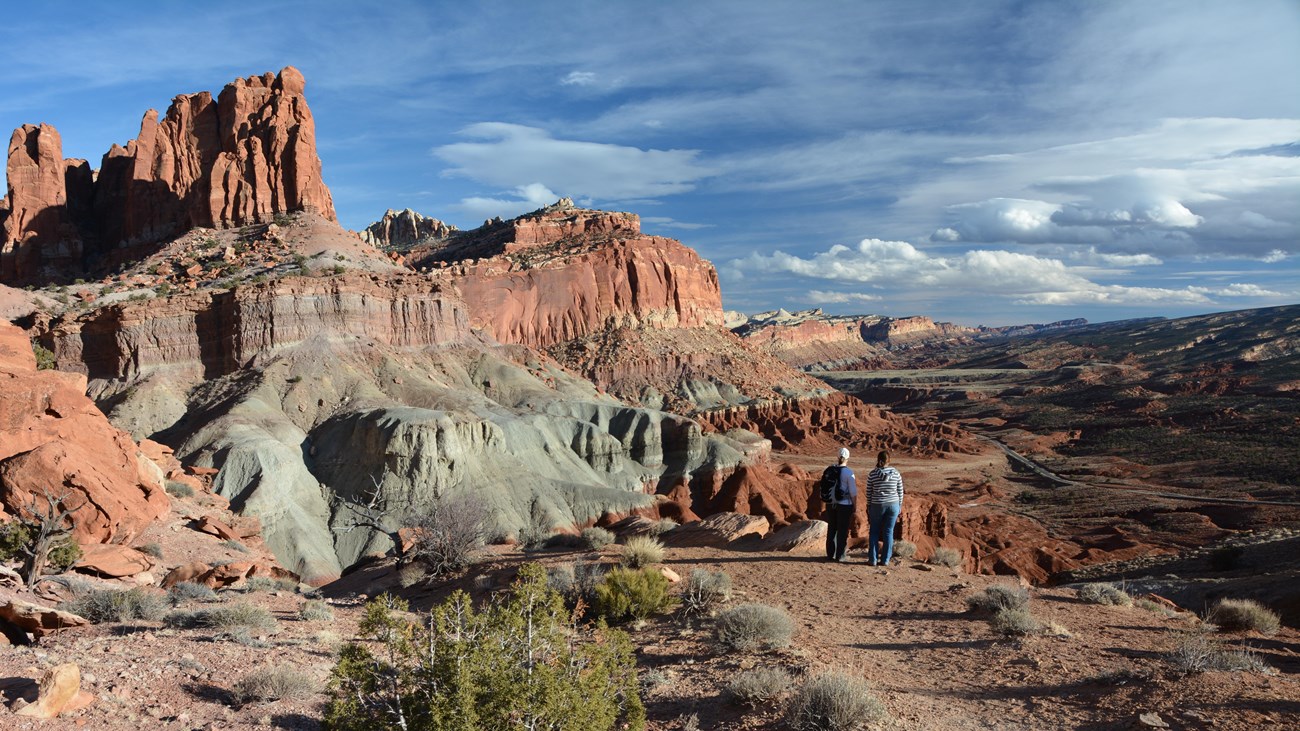 Two hikers stand at the edge of a cliff, looking at a panoramic view of colorful rock cliffs.