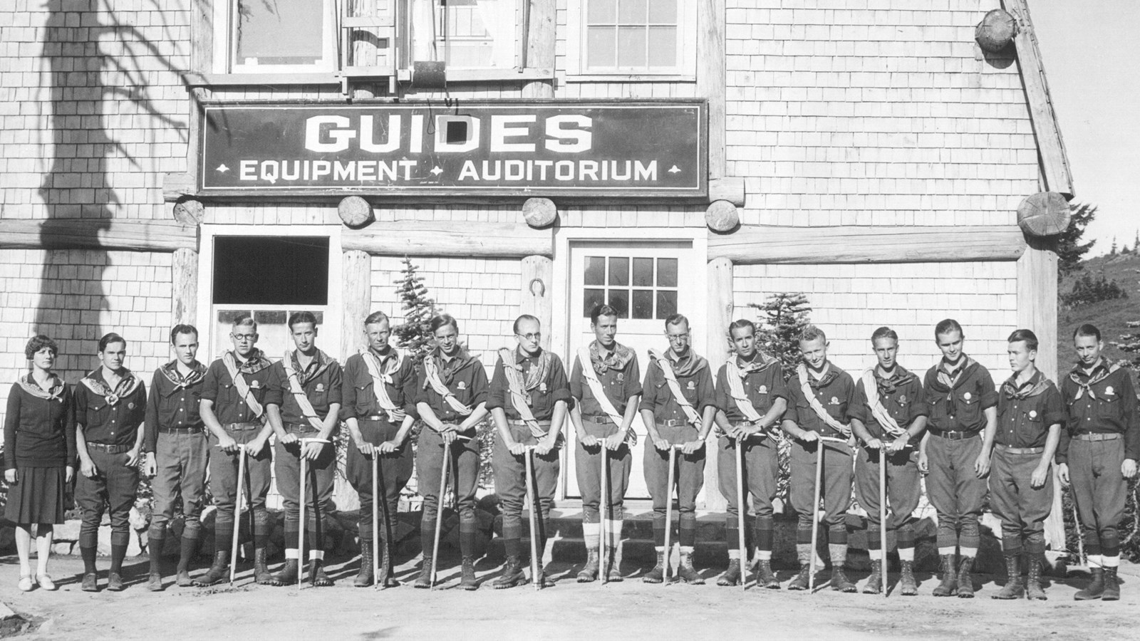 A black and white photo of guides with ropes and ice axes lined up in front of wooden building