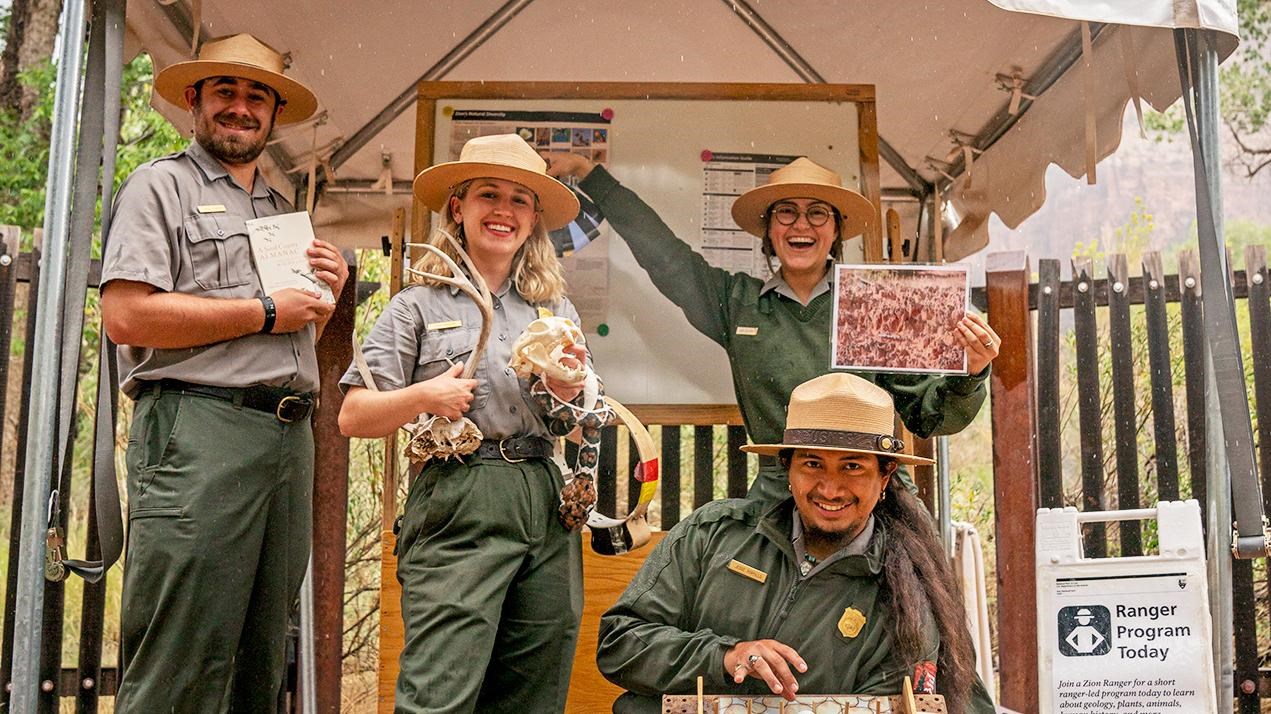 Four NPS rangers stand under a shade structure holding various props.
