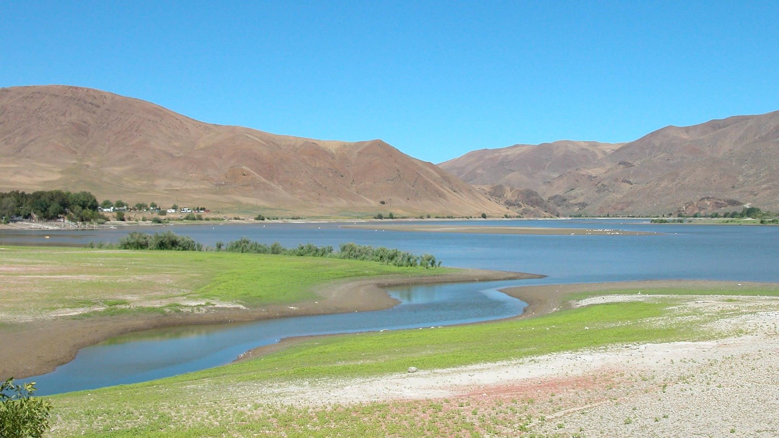 A calm, wide river, lightly banked with grass and distant desert mountains.