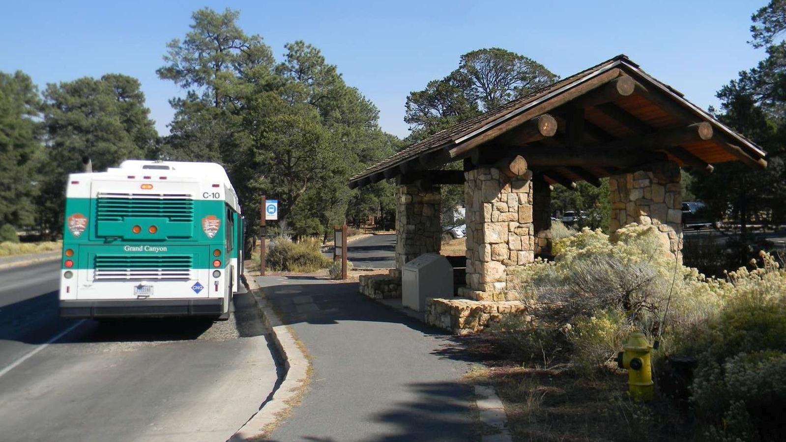 A large white and green bus sits next to a stone shelter with a wooden roof.