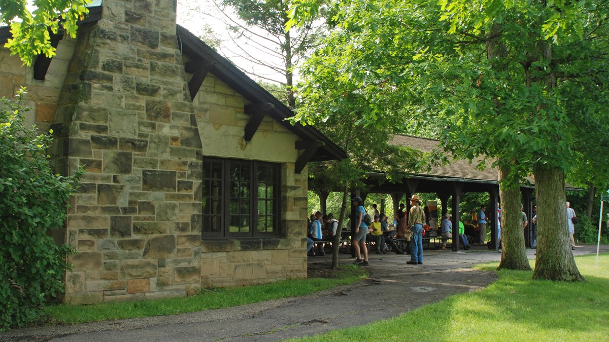 A crowd gathered around picnic tables under a rustic shelter made of sandstone and dark brown wood.