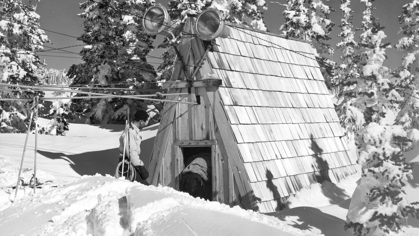 A steep A-frame roof building sits in deep snow surrounded by trees