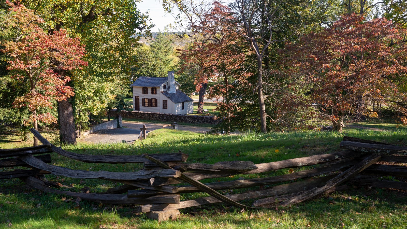 View from atop a hill, looking down on a gravel road and small white house through trees in fall.