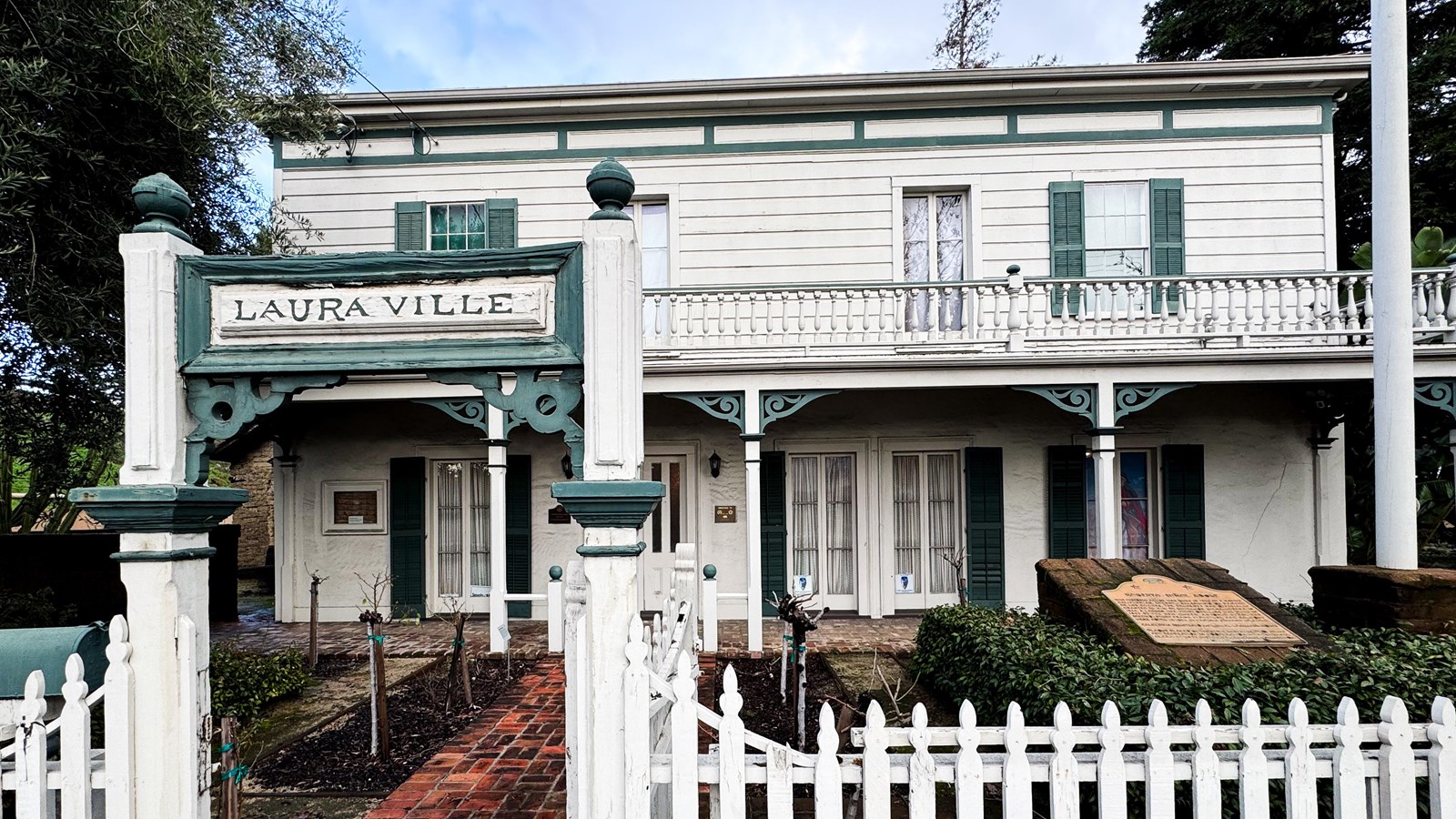 A two-story white house with green trim has a white picket fence with a sign that says Laura Ville