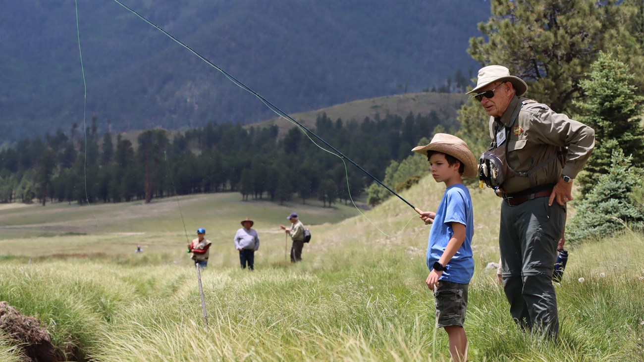 A boy wearing a cowboy hat flyfishes a narrow stream in a montane grassland.
