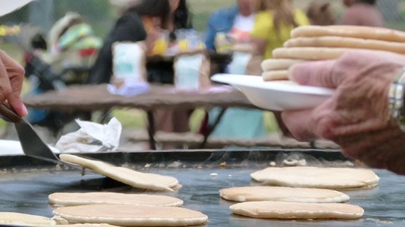 Close-up of pancakes on a griddle being served. People eat at picnic tables in the background.