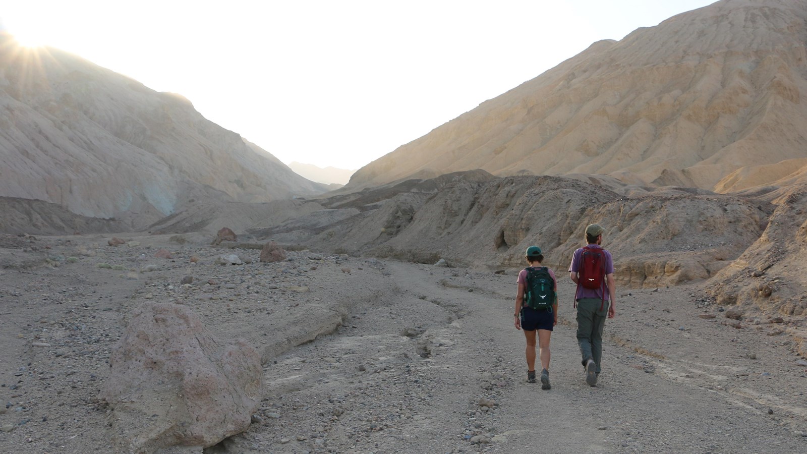 Two hikers walk up a wash with their backs to the camera, while the sun rises over the canyon wall.