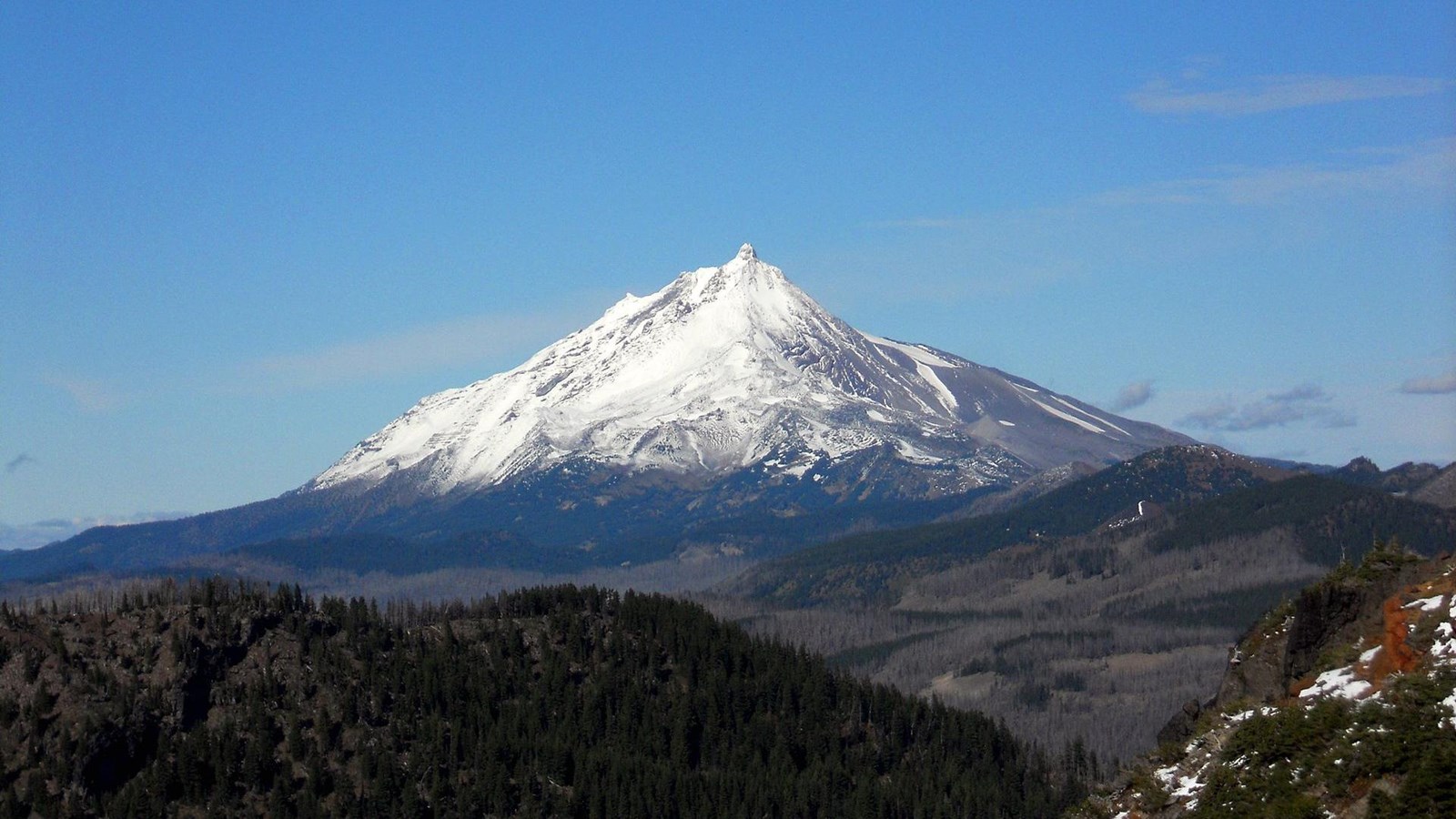 Mount Jefferson’s white peak stands tall against a bright blue sky, with tree-covered hills in the f