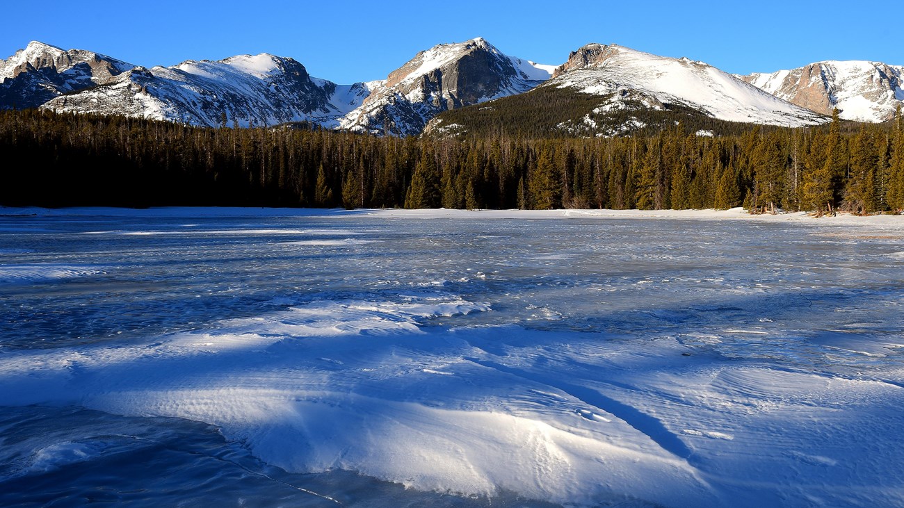 The lake is frozen with a layer of snow on top. Mountains in the distance are covered with snow