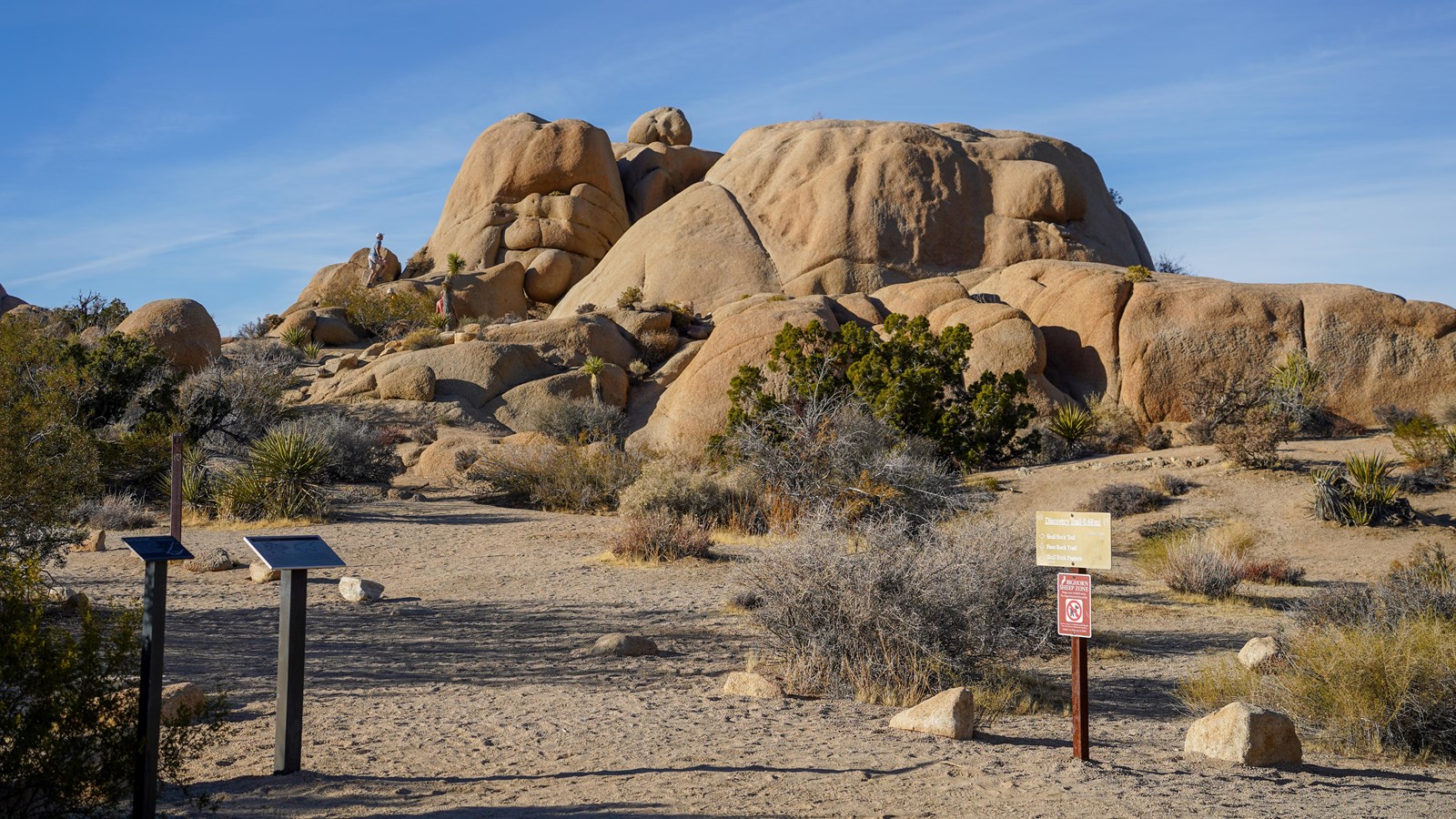 Interpretive signs stand on either side of a dirt path leading to a giant boulder pile and plants.