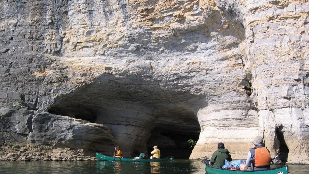 green canoes on river in front of grey cliff with large hole at river level