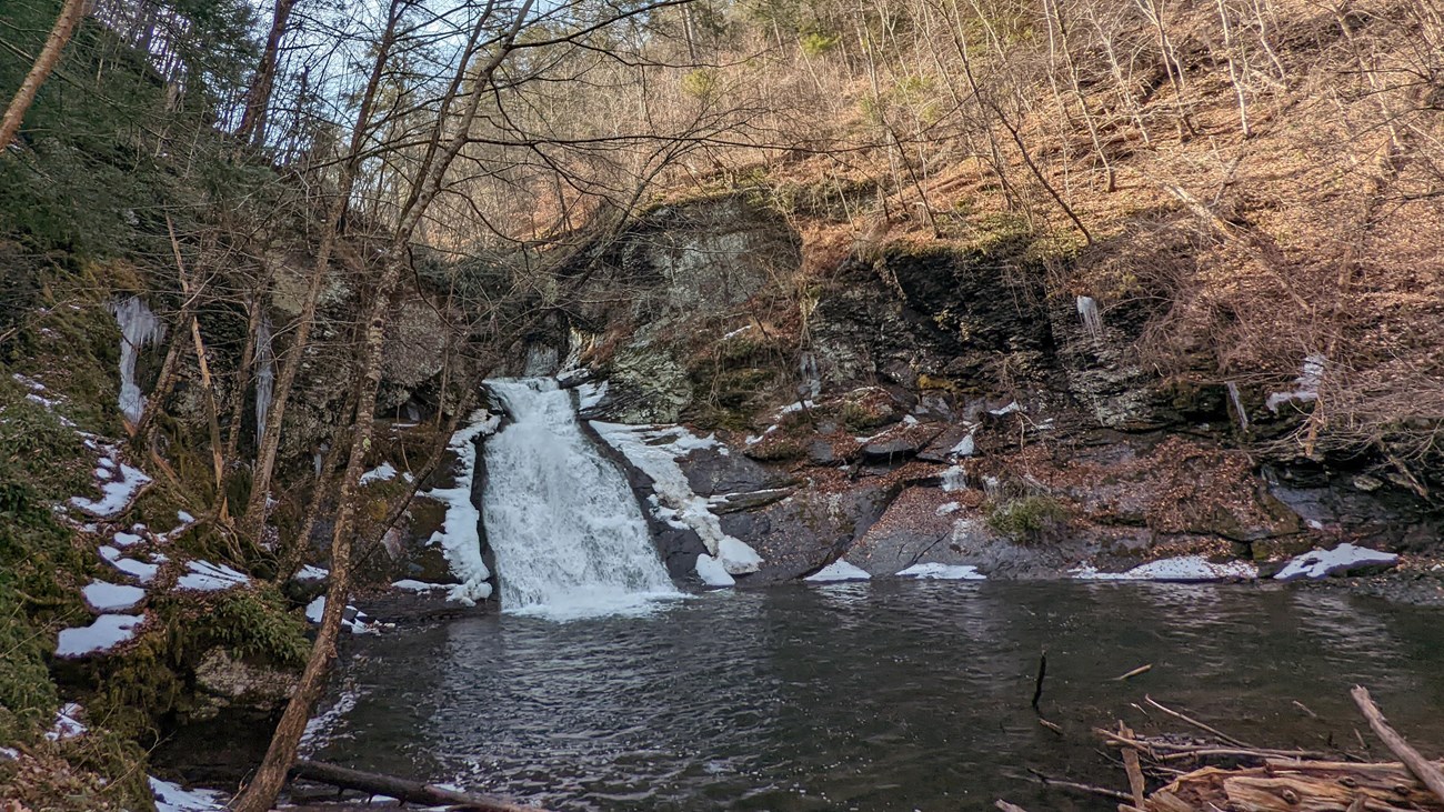 A small, tiered waterfall and pool with a small amount of snow around it.