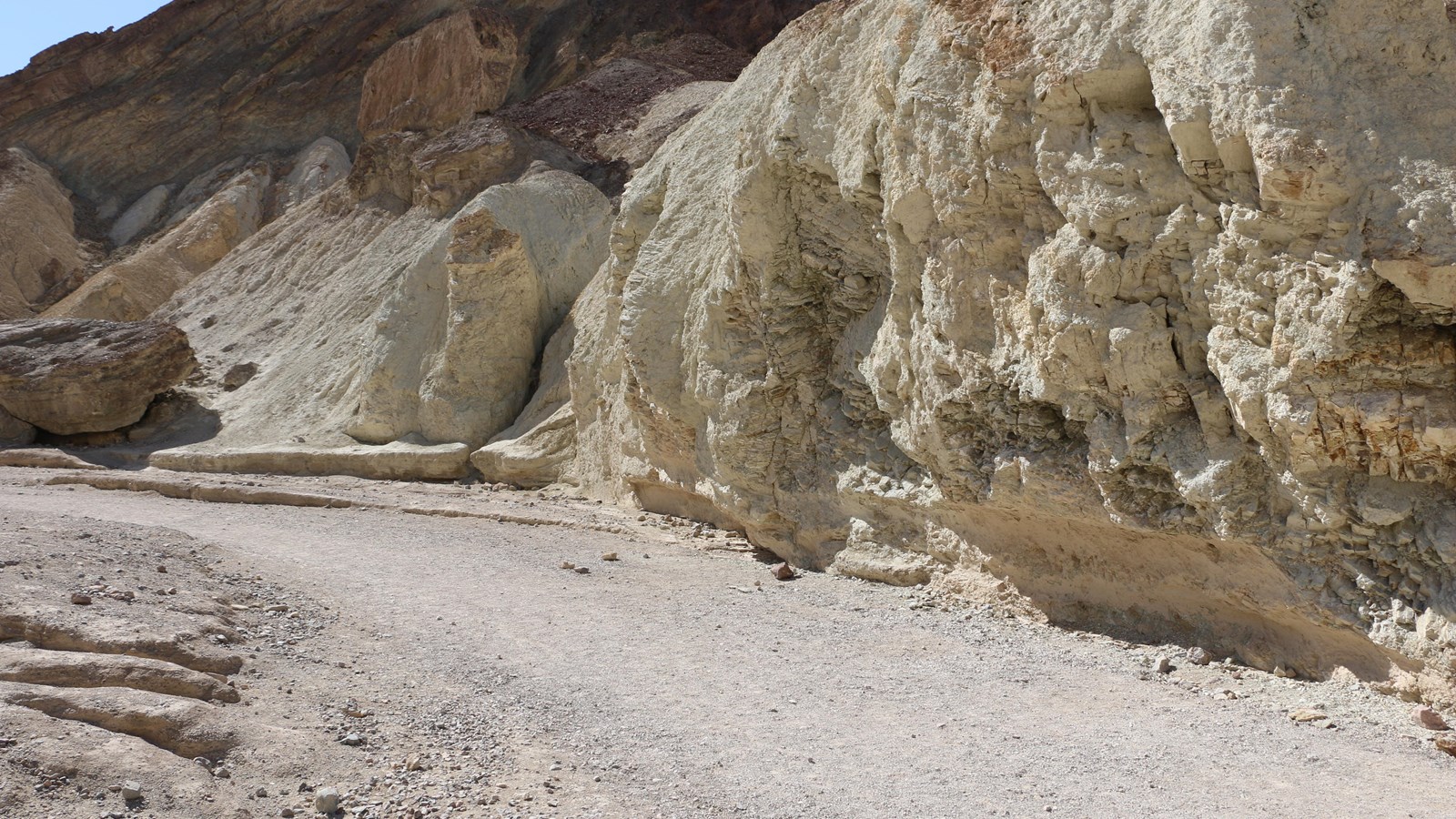 A wash runs at the base of a bare, tan-colored wall with a darker/higher canyon wall behind it.