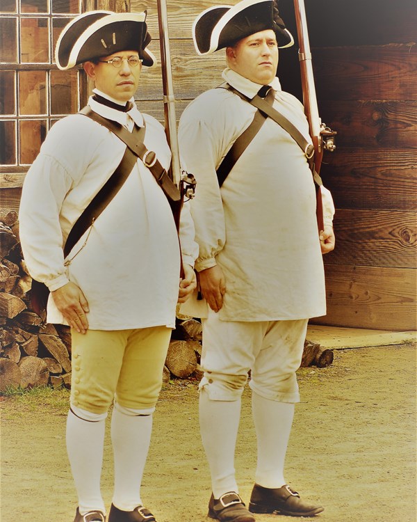 An image of two men standing next to each other. They are ridged and hold muskets at the shoulder.