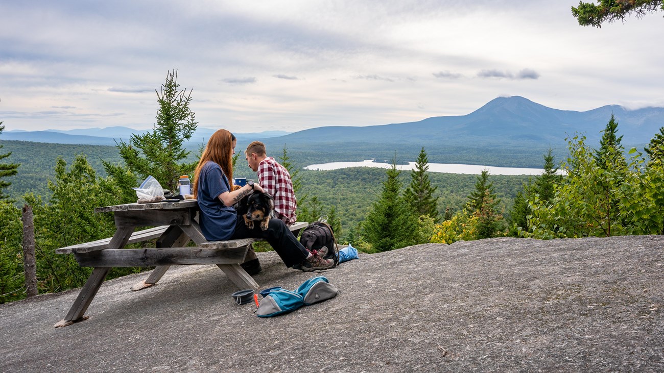 Two hikers and their dog on a leash have a picnic with a view on top of Barnard Mountain.
