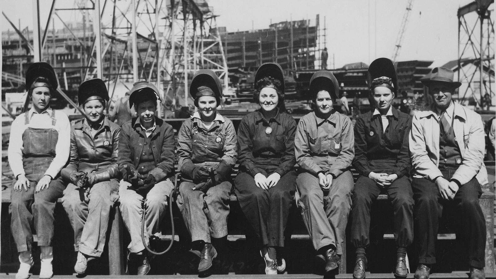women in coveralls and one man in trench coat sitting in a line on a girder.
