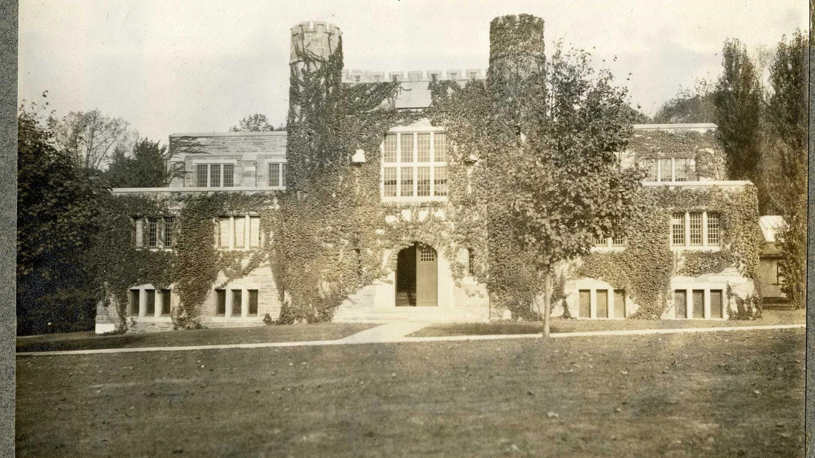 Black and white of building with vines covering it with grass in front and paths leading to it. 