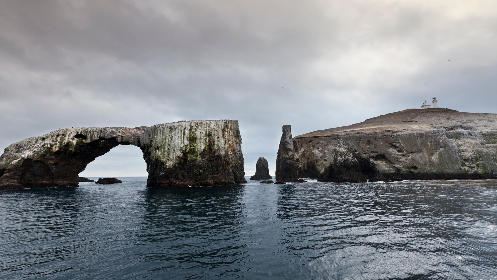 A cliff island with an lighthouse near the top and an arched rock off shore.