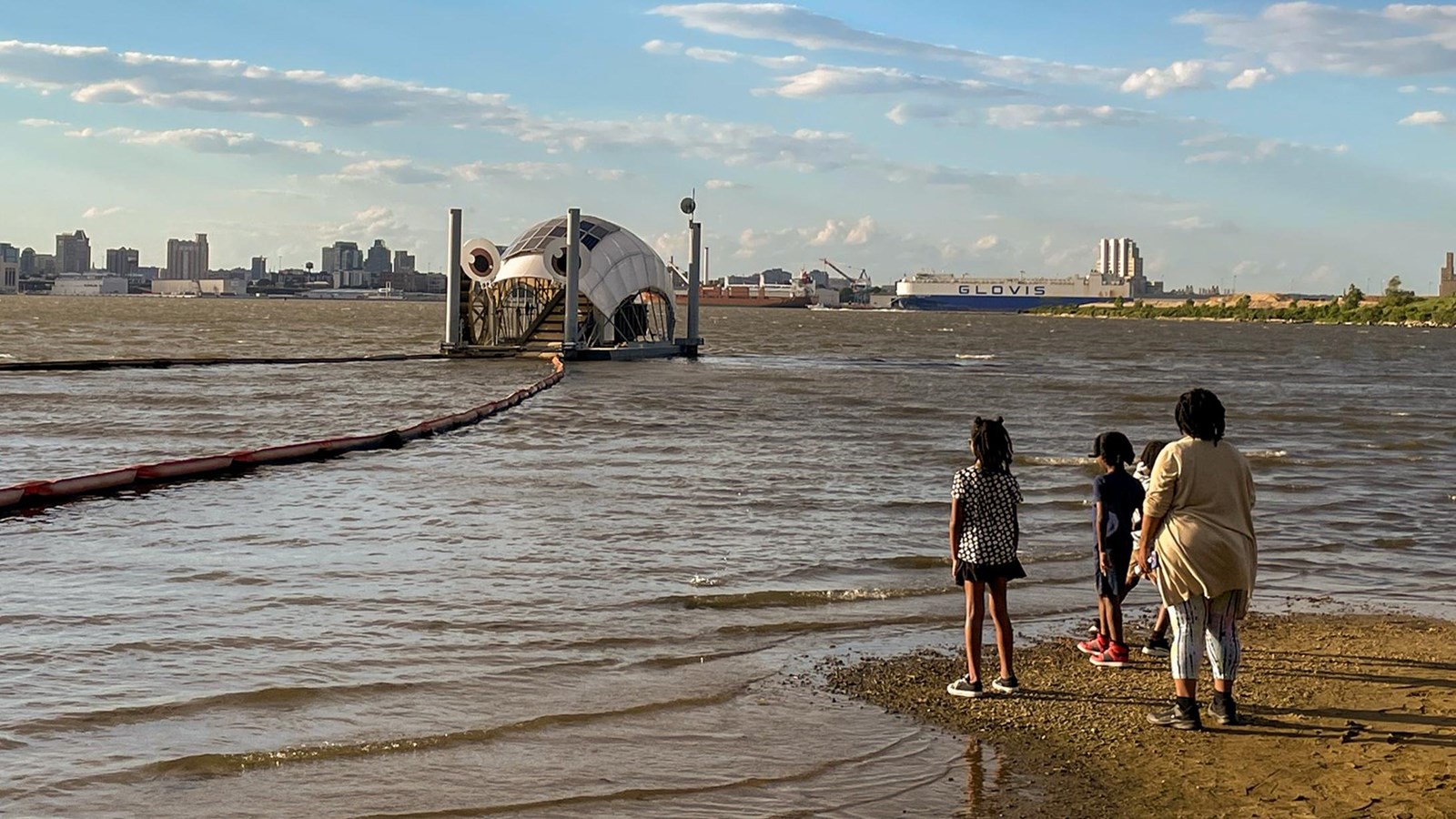A family looks out at a floating dome structure on a river, with a city skyline in the background.