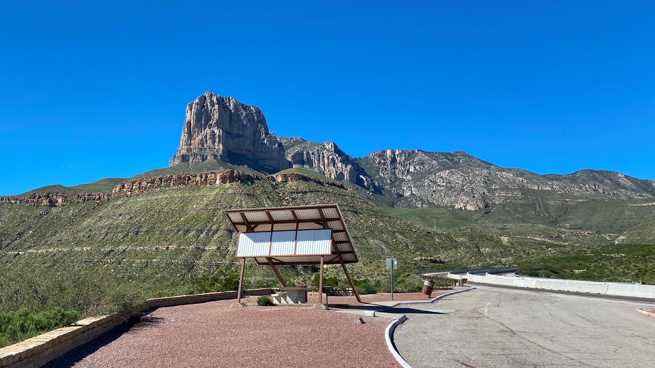 A roadside picnic shelter stands below a prominent mountain peak. 