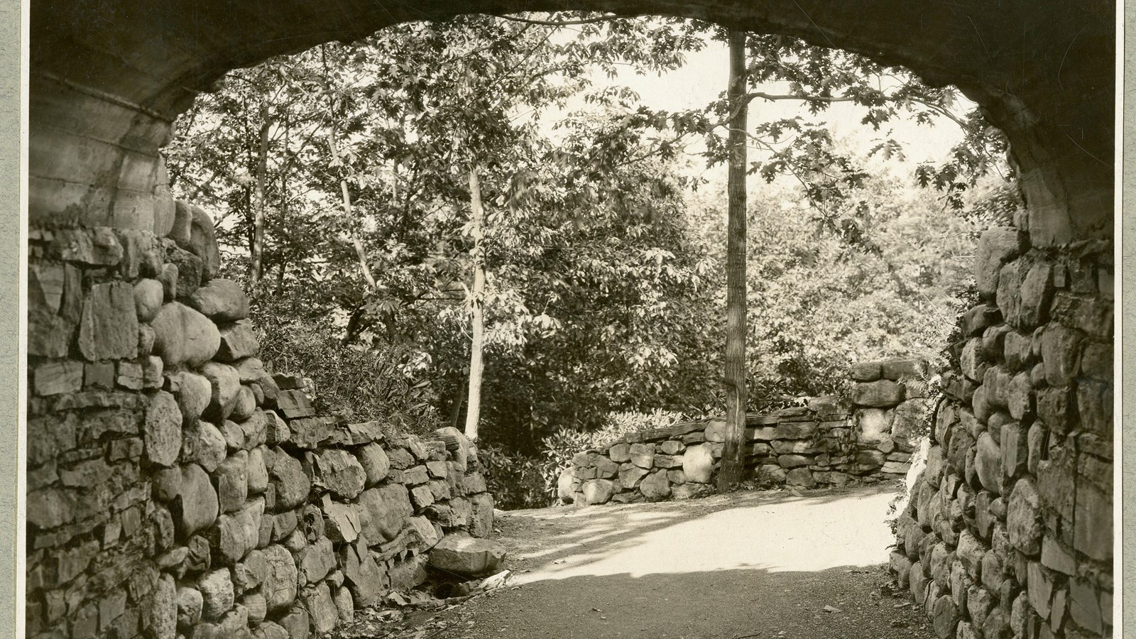 Black and white underneath stone bridge looking at stone wall and trees