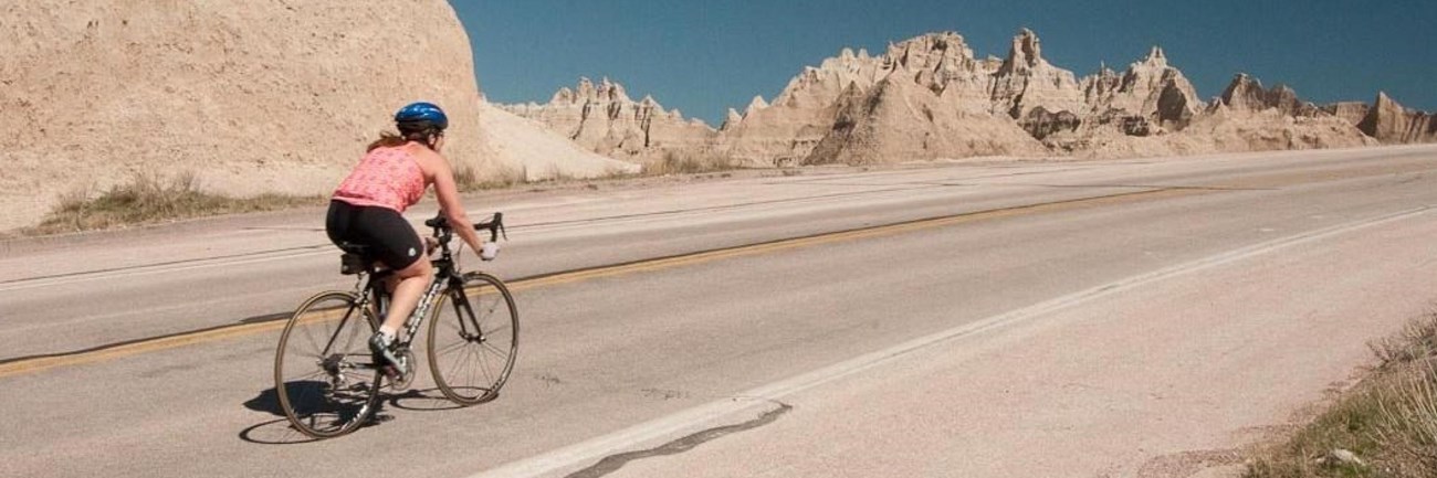a woman bikes up a paved road with jagged badlands spires in the background