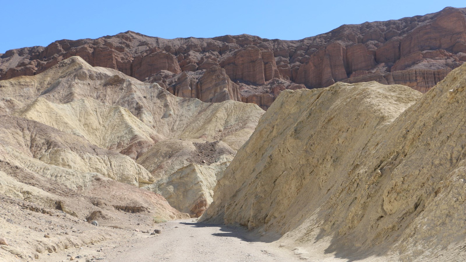 A tall red cliff with eroding pillars towers behind bare golden hills with a gravel wash in front.