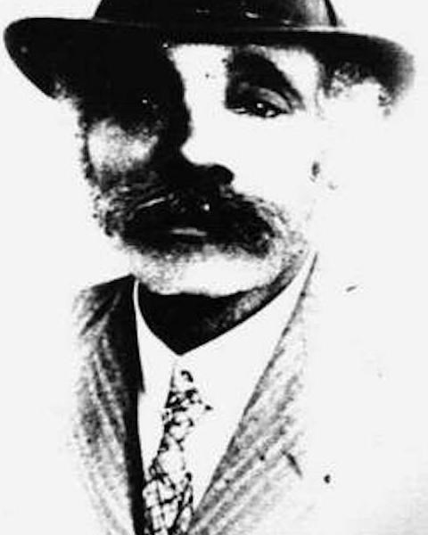 A black and white portrait of Mr. Fletcher. He wears a brimmed hat, suit and tie, and has a mustache
