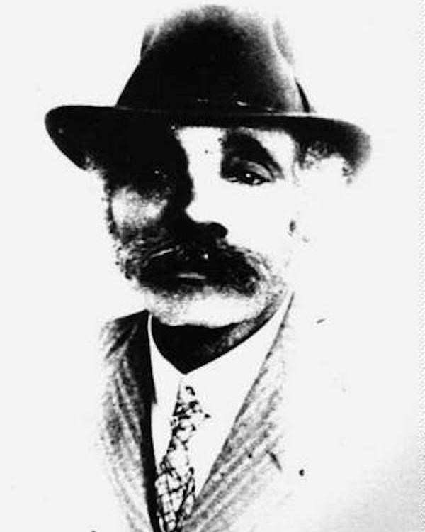 A black and white portrait of Mr. Fletcher. He wears a brimmed hat, suit and tie, and has a mustache