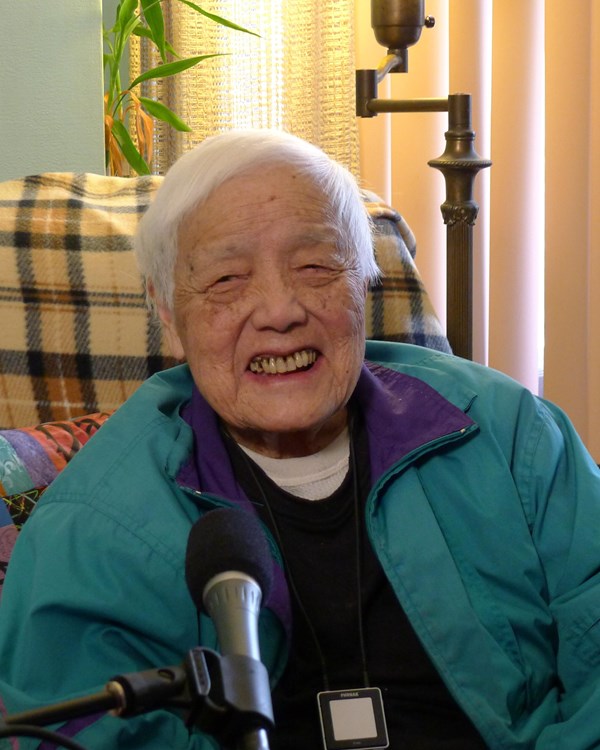 Elderly woman wearing a green jacket and smiling in front of a microphone.