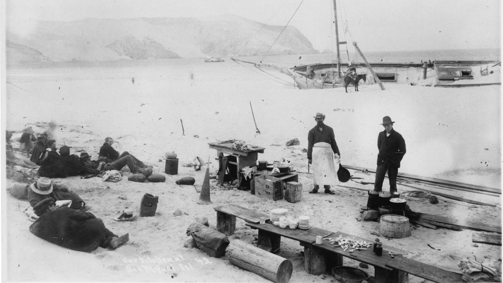 Visitors cooking on beach with shipwreck behind them.