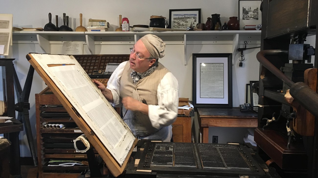 man in historical vest and cap with glasses looking at a printed page from a press.