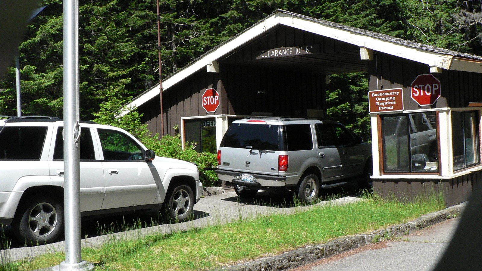 Vehicles wait in line at a small entrance station with two booths covered by a shared angled roof. 