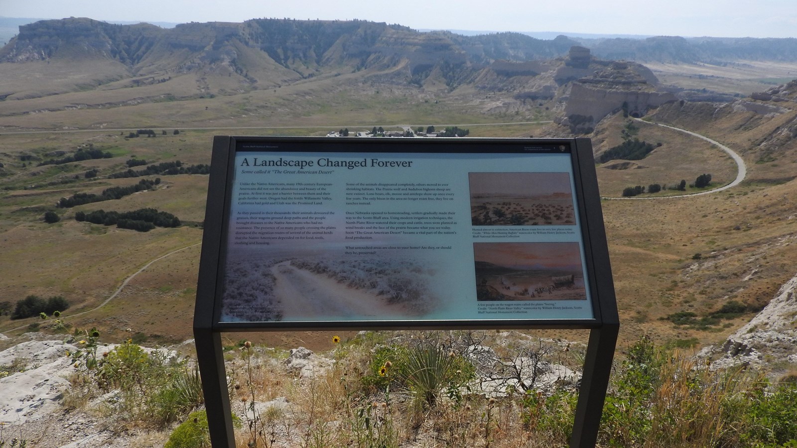 An interpretive panel sits at the edge of a steep cliff overlooking sandstone bluffs. 