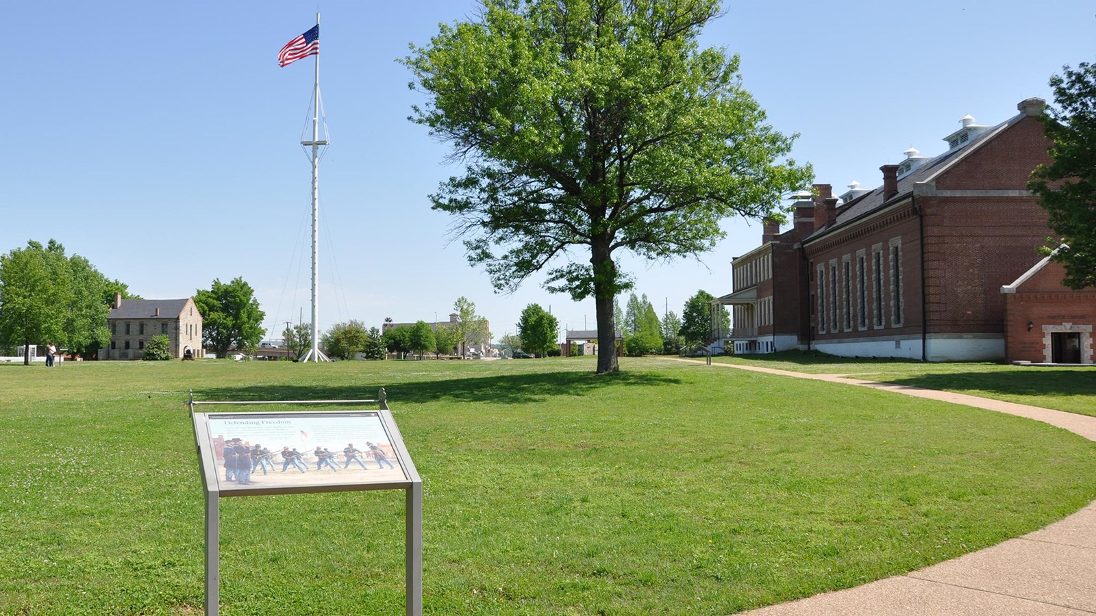 Wayside at the edge of green field, parade ground. Buildings can be seen in the background.