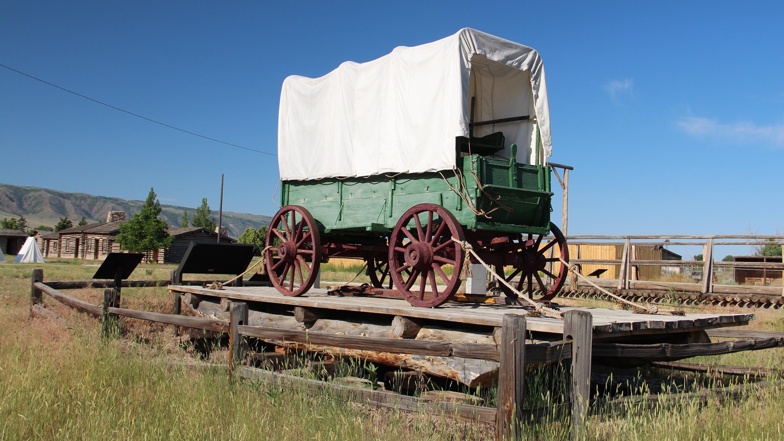 Green wagon secured on a plank ferry with fence, exhibits, and log buildings in view.