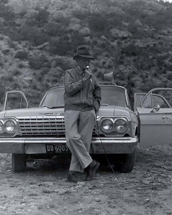 A man speaks into a microphone while leaning against a car